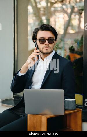 Junge gut aussehende seriöse bärtige Brünette Mann in weißem Hemd, klassische Jacke und Sonnenbrille im Gespräch auf Handy nachdenklich Blick beiseite während des Sitzens Stockfoto