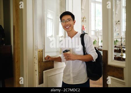 Junge asiatische männliche Student in Brillen mit Rucksack und Kaffee zu gehen Öffnung Tür Bibliothek in der Universität Stockfoto