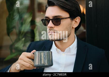 Junge gut aussehende bärtige Brünette Mann in weißem Hemd, klassische Jacke und Sonnenbrille nachdenklich Blick beiseite, während die Tasse Kaffee in den Händen halten Stockfoto