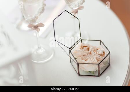 Eheringe in der Glasbox gefüllt mit Rosen auf dem weißen Tisch stehen. Hochzeitsaccessoires Stockfoto