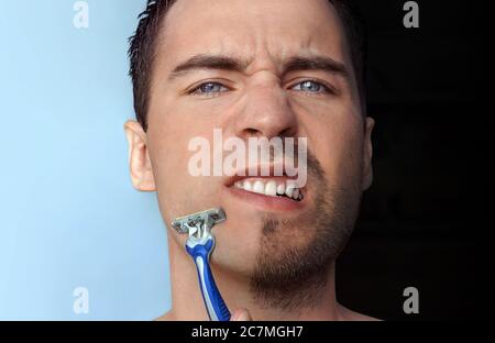 Der Mensch rasiert sein Gesicht ohne Creme oder Schaum und erlebt Schmerzen und Leiden. Halb Gesicht rasiert halb überwuchert mit einem dicken Bart Nahaufnahme. Copy Space Stockfoto