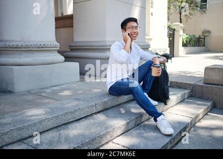 Junge lächelnde asiatische männliche Student in Brillen mit Rucksack glücklich reden auf Handy sitzen auf Treppen in der Nähe der Universität Stockfoto
