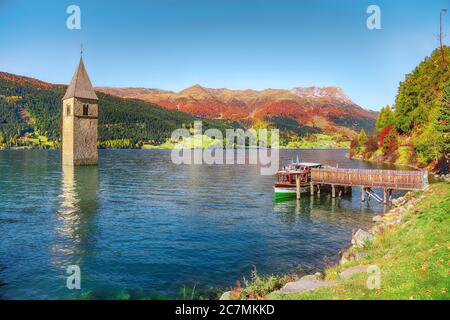 Fantastische Herbst Seenlandschaft mit einem Schiff in der Nähe der Pier und untergetauchten Glockenturm im See Reschen. Lage: Graun im Vinschgau Dorf, Reschensee Stockfoto