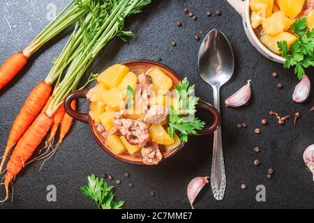 Kartoffelschmorbraten, gebacken mit Fleisch, Schweinefleisch und Petersilie auf einem tiefen Teller neben einem Gewürz numerisch Karotte auf einem schwarzen Tisch. Flach legen, Kopierraum, Draufsicht Stockfoto