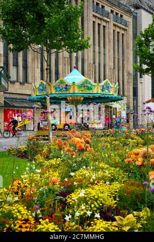 Nostalgisches Karussell vor dem historischen Gebäude des Kaufhofs in Düsseldorf, mit bunten Sommerblumen im Vordergrund. Stockfoto