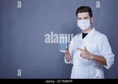 Männlicher Arzt mit weißer medizinischer Robe und Gesichtsmaske Zeigt ein Smartphone an Stockfoto