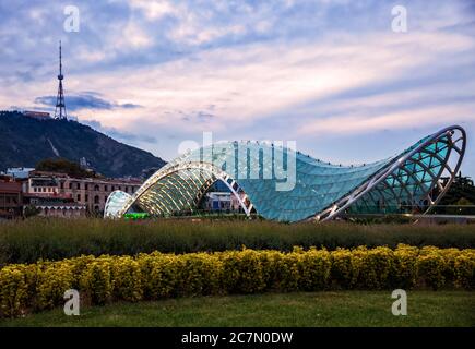 Brücke des Friedens vor dem Hintergrund des Berges Mtasminda und des Fernsehturms. Tiflis Stadtbild am Abend bei Sonnenuntergang Stockfoto