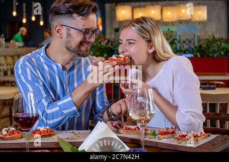 Happy junge romantische Paar in der Liebe Fütterung einander mit Bruschetta beim Abendessen in einem schönen schicken Restaurant. Stockfoto