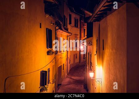 Chiusi, Italien leere Straße schmale Gasse und Katze auf Balkon Fenster in kleinen historischen Dorf in der Toskana während der Sommernacht mit niemand orange yel Stockfoto