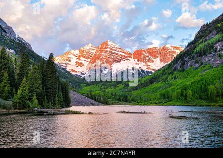 Kastanienbraune Glocken in Aspen, Colorado mit Wolken in blauem Himmel bei Sonnenaufgang und felsiger Bergspitze See Reflexion im Sommer mit rotem Sonnenlicht und grünen Fores Stockfoto