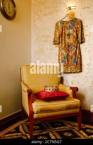 Eine goldene Krone mit roten und blauen Edelsteinen, auf einem roten Kissen auf einem Stuhl im Innenraum des Zimmers. An der Wand hängt ein goldenes Kleid. Stockfoto