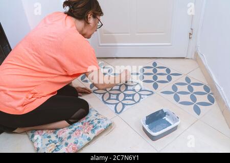 Zu Hause bleiben und Heimwerker-Konzept: Junge kaukasische Frau mit orangefarbenem T-Shirt malt Fliesen von ihrem Haus. Stockfoto