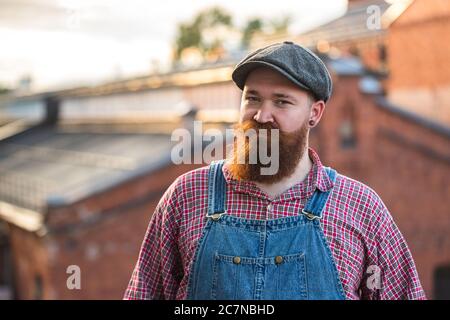 Portrait eines brutalen bärtigen, stylischen Handwerkers mit blauen Overalls, kariertem Hemd und Mütze im Vintage-Stil der Mitte des 20. Jahrhunderts, mit Blick auf die Kamera Stockfoto