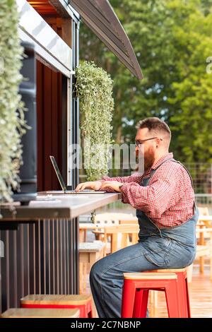 Portrait eines brutalen bärtigen Hipster-Mannes, der als Freiberufler blaue Overalls und kariertes Hemd trägt, der auf einem Laptop im Café/Restaurant im Freien sitzt. Seite Stockfoto