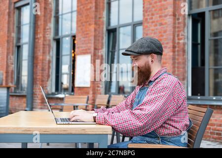 Portrait eines brutalen bärtigen Hipster-Mannes, der als Freiberufler blaue Overalls und kariertes Hemd trägt und auf einem Laptop im Café/Restaurant im Freien sitzt. Seite Stockfoto