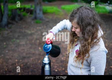 Frau hält Tee Wasserkessel aus der Feuerstelle am Campingplatz mit Wasser in Behälter am Abend oder Morgen mit Fäustling Stockfoto