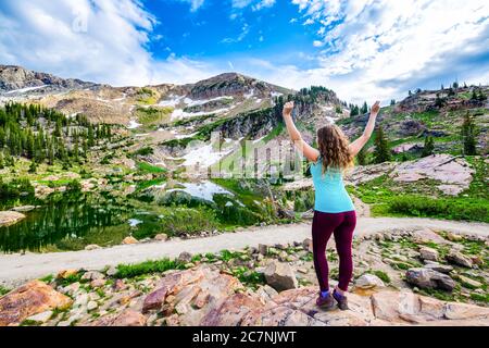 Albion Basin, Utah Sommer mit glücklicher Frau zurück stehend Blick auf die Ansicht Reflexion des Wassers auf Cecret Lake in Wasatch Berge mit Felsen, Schnee und Stockfoto