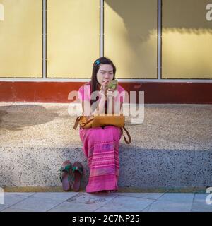Eine junge Frau sitzt auf einer Bordsteinkante und schaut auf ihr Handy in der Shwedagon Pagode in Yangon, Myanmar Stockfoto