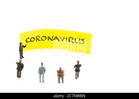 Treffen mit Miniaturfiguren, die als Geschäftsleute um Post-it-Notiz mit handgeschriebener Coronavirus-Nachricht im Hintergrund stehen, minimalistisch Stockfoto
