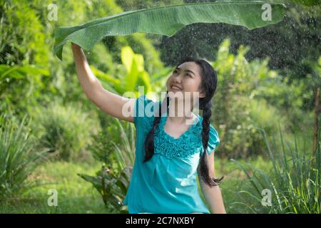 Porträt einer jungen Asienfrau mit schwarzem Haar, die ein Bananenblatt im Regen auf dem grünen Gartenhintergrund hält Stockfoto