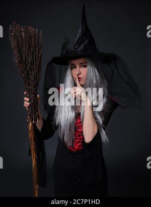 Geheimnisvolle Frau in schwarzem Hexen halloween Kostüm und Hut mit Besen zeigt shh Zeichen über schwarzem Hintergrund Stockfoto