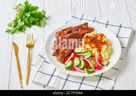Schweinebrippen in einem Bier serviert mit Kartoffelpüree und Gemüsesalat mit Tomaten, Gurken, roten Zwiebeln, Petersilie auf einem weißen Teller mit goldenem Cu Stockfoto