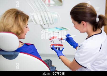 Attraktive weibliche Zahnarzt hält künstliche Kiefer und zeigt fröhlich reife Frau Patientin, wie man Zähne putzt Stockfoto