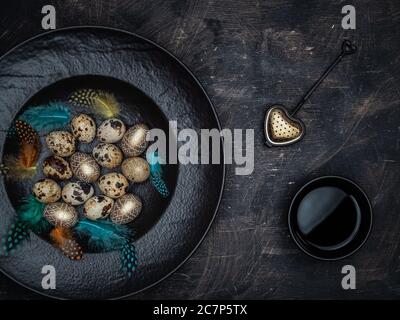 Natürliche Wachteleier, goldfarbene Eier und bunte Federn in schwarzer Schale. Tasse schwarzen Tee und Tee Sieb. Stockfoto