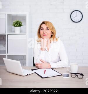 Fröhliche reife Geschäftsfrau sitzt in modernen Büro und träumt von etwas Stockfoto