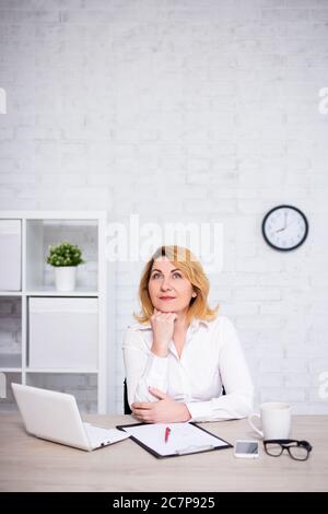 Fröhliche reife Geschäftsfrau sitzt im Büro und träumt von etwas - kopieren Raum über weißen Ziegelwand Stockfoto