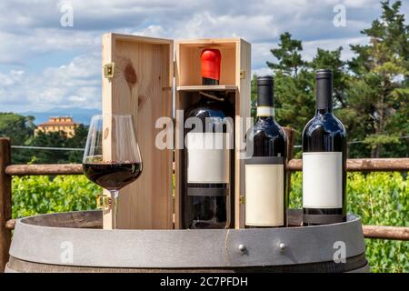 Drei Flaschen, darunter ein Magnum in der Holzkiste und ein Glas Rotwein auf einem Holzfass mit der toskanischen Landschaft im Hintergrund, Italien, auf Stockfoto