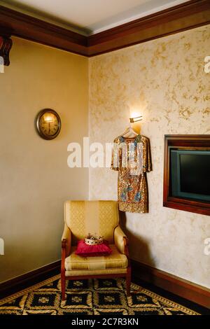 Eine goldene Krone mit roten und blauen Edelsteinen, auf einem roten Kissen auf einem Stuhl im Innenraum des Zimmers. An der Wand hängt ein goldenes Kleid. Stockfoto