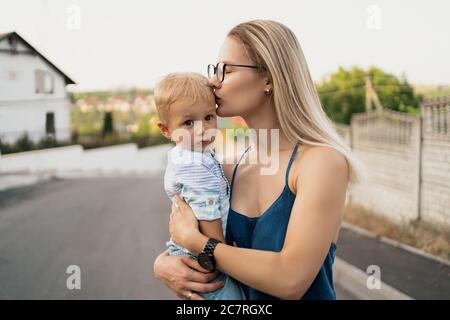 Frau trägt ihren Sohn und küsst sich auf seiner Stirn. Niedlicher kleiner Junge in den Armen seiner Mutter. Stockfoto