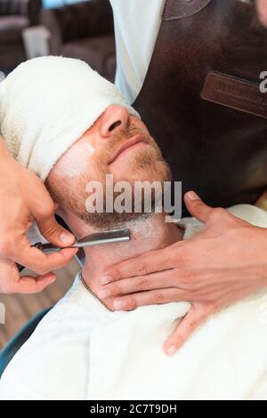 Vertikale Aufnahme eines Friseurs rasieren den Bart eines Junger Kunde in einem Handtuch mit Klinge Stockfoto