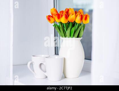 Nahaufnahme von schönen orangefarbenen künstlichen Tulpen und zwei Tassen auf einer Fensterbank Stockfoto