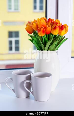 Nahaufnahme von schönen orangen Kunstblumen und zwei Tassen auf einer Fensterbank Stockfoto