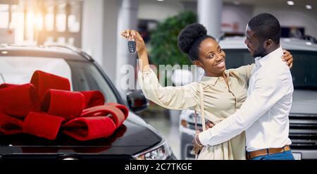 Abenteuer erwartet. Schöne glückliche junge afrikanische Paar umarmt hält die Schlüssel zu ihrem neuen Auto lächelnd freudig auf den Händler. Verliebt Stockfoto