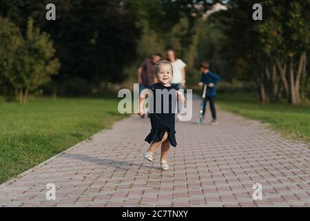 Schönes kleines Mädchen läuft glücklich, während ihre Mutter, Bruder, Vater ihr folgen, während sie im Park spazieren Stockfoto