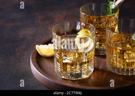 Drei Gläser kalter Whiskey auf Felsen mit Rosmarin serviert, Zitronenschalen auf braunem Hintergrund. Speicherplatz kopieren. Stockfoto