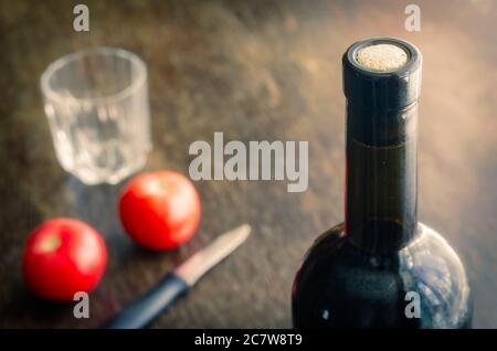 Versiegelte Flasche Rotwein auf Holztisch mit reifen Tomaten. Volle Flasche Wein mit Kork, zwei roten Tomaten, Messer und ein leeres Glas. Konzentrieren Sie sich auf die Stockfoto