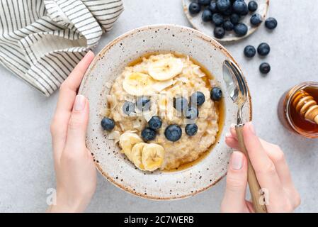 Haferflocken Haferbrei mit Banane, Heidelbeeren und Honig. Weibliche Hände halten Schüssel Haferbrei Haferbrei auf grauem Beton Hintergrund, Tabelle Draufsicht Stockfoto