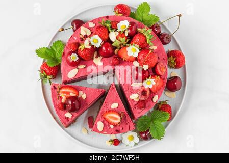 Beerenkochkuchen mit frischen Erdbeeren, Kirschen und Blumen auf weißem Hintergrund in Scheiben geschnitten. Draufsicht. Gesundes veganes Dessert Stockfoto