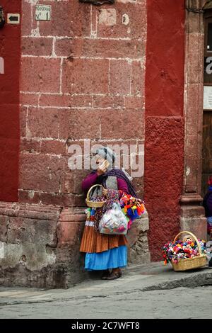 Eine indigene Frau spricht auf ihrem Handy, während sie handgemachtes Kunsthandwerk auf der Plaza Allende in San Miguel de Allende, Guanajuato, Mexiko verkauft. Stockfoto