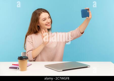 Fröhliche Frau, Büroangestellte oder Blogger, am Arbeitsplatz sitzend und Selfie per Handy, Gespräch auf Video-Anruf mit hallo Geste, willkommenin Stockfoto