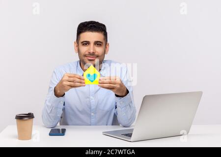 Portrait von glücklichen Immobilienmakler Mann sitzen in Büro-Arbeitsplatz und halten Papierhaus mit Herz im Inneren, lächelnd freundlich, Werbung von purc Stockfoto