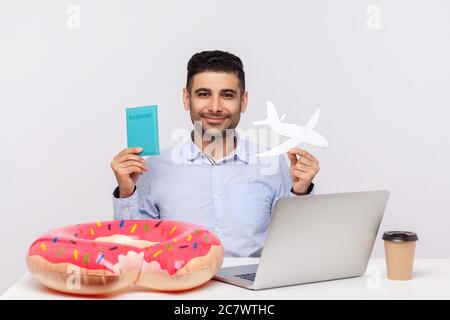 Fröhlicher Mann, der Papierflugzeug und Reisepass in den Händen hält, am Büroarbeitsplatz mit Gummiring am Schreibtisch sitzt, Reiseurlaub plant, Wochenende Tri Stockfoto