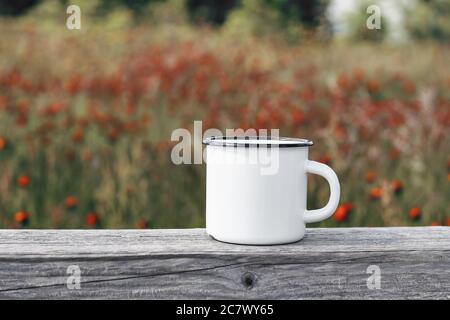 Nahaufnahme von Metallbecher auf alten Holztisch, Brett mit unbelauchten blühenden Bergwiese. Tee im Freien, Kaffee Zeit. Modell aus weißem Emaille-Becher Stockfoto