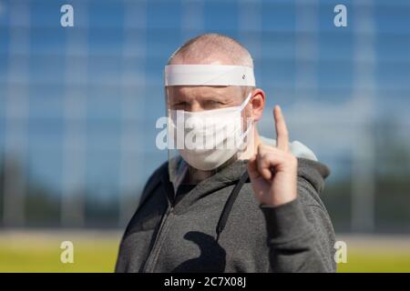 Erwachsene Mann posiert in einem schützenden transparenten Visier und einer weißen Maske auf seinem Gesicht im Freien an einem sonnigen Tag Stockfoto