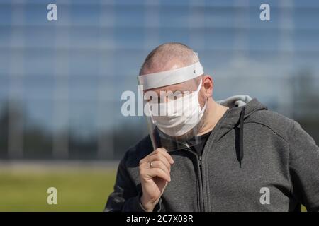 Erwachsene Mann posiert in einem schützenden transparenten Visier und einer weißen Maske auf seinem Gesicht im Freien an einem sonnigen Tag Stockfoto