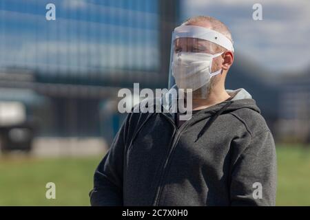 Erwachsene Mann posiert in einem schützenden transparenten Visier und einer weißen Maske auf seinem Gesicht im Freien an einem sonnigen Tag Stockfoto